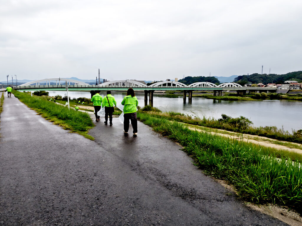 alt="高橋建設社員が益田市の高津川左岸でペットボトルや空き缶を回収する清掃活動の様子"