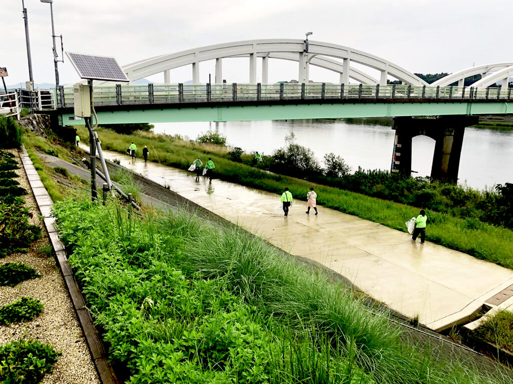 alt="高橋建設社員が益田市の高津川左岸でペットボトルや空き缶を回収する清掃活動の様子"