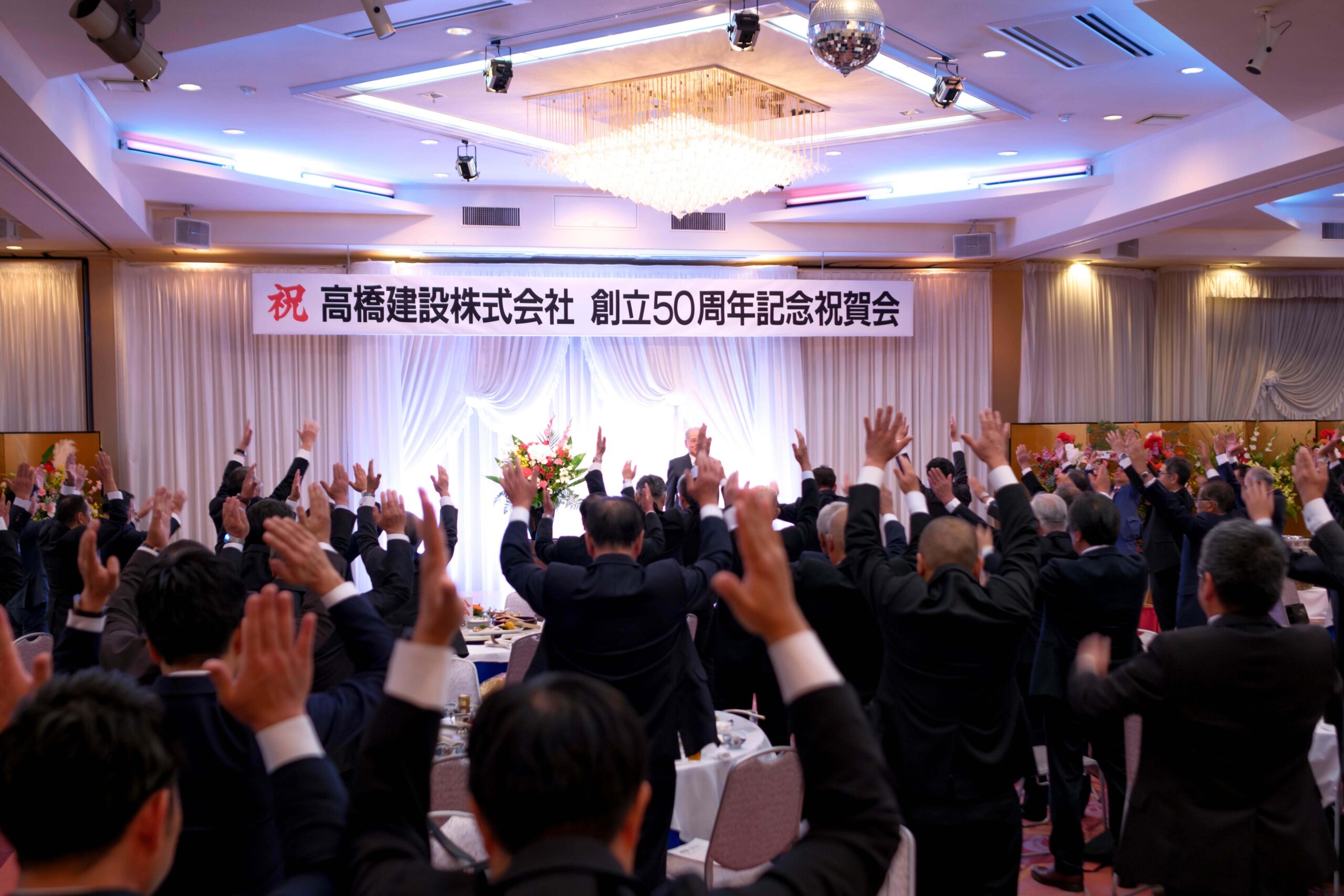alt=“高橋建設 創業50周年祝賀会で万歳三唱をする万歳三唱益田地区防衛協力会会長、益田酒類食品(株) 代表取締役島田憲郷様”